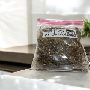 May include: A clear plastic bag with a pink zipper closure, filled with dried plant material. The bag has handwritten text that reads "Fey ti chien Scambe" and "Date Contents". The bag is sitting on a white countertop.