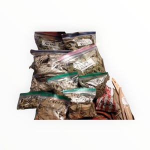 May include: A collection of clear plastic bags filled with dried herbs and leaves. The bags are labeled with handwritten text. Some bags have red or blue zippers. There are also bundles of wooden sticks and a small red and white striped bag.