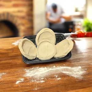 Könnte beinhalten: Ein schwarzes Plastiktablett enthält fünf rohe, beige Pizzateiglinge. Der Teig liegt auf einem Holztisch, der mit Mehl bestäubt ist. Im Hintergrund sind ein Ziegelofen und eine Person leicht unscharf.