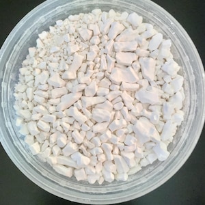 Puede incluir: Un recipiente de plástico transparente lleno de trozos irregulares de color blanquecino. Las piezas parecen ser un material sólido, posiblemente un polvo o una sustancia granular. El recipiente está sobre un fondo oscuro.