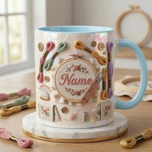 Puede incluir: Una taza de cer&aacute;mica blanca con interior y asa azul claro, decorada con adornos tem&aacute;ticos de bordado. La taza presenta carretes de hilo, botones y un peque&ntilde;o bastidor de bordado con el nombre "Name" en cursiva. La taza se apoya en un posavasos de m&aacute;rmol.