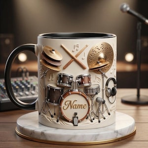 Könnte beinhalten: Cremefarbene Keramiktasse mit einem detaillierten Schlagzeug-Design mit Becken, Trommeln und Drumsticks. Die Tasse hat einen schwarzen Henkel und Rand und steht auf einem Marmoruntersetzer. Das Wort "Name" ist auf der Trommel zu sehen.