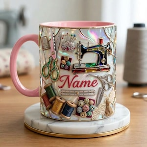 Könnte beinhalten: Eine dekorative Tasse mit rosa Griff und Rand, verziert mit Näh-Elementen. Das Design umfasst eine Nähmaschine, Scheren, Garnrollen und Knöpfe. Das Wort "Name" ist in roten Buchstaben dargestellt.