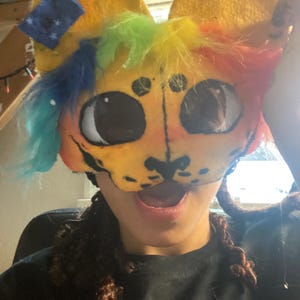 May include: A colorful, handmade animal mask with large, expressive eyes and a smiling mouth. The mask features yellow fur, rainbow-colored accents, and a small blue patch with stars. The mask is designed to be worn.