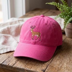 Könnte beinhalten: Eine leuchtend pinke Baseballkappe mit einem gestickten braunen Hirsch mit dunklem Geweih in der Mitte des Vorderteils. Die Kappe hat einen gebogenen Schirm, einen Knopf an der Oberseite und genähte Ösen zur Belüftung aus festem Stoff.