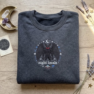 May include: A charcoal grey crewneck sweatshirt featuring an embroidered design of a winged creature with red eyes, a crescent moon, stars, and a small flying saucer. The text "night locals" is stitched in white below the central figure.