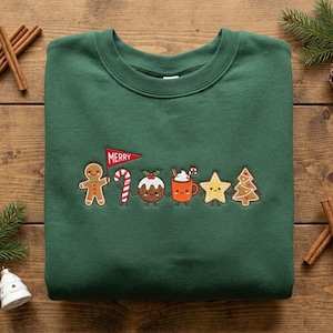 May include: A dark green crewneck sweatshirt featuring an embroidered design of six holiday characters holding hands: a gingerbread person holding a red pennant flag that says MERRY, a candy cane, a pudding, a cocoa mug, a star, and a tree.