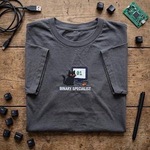 May include: A charcoal gray t-shirt featuring an embroidered graphic of a black cat sitting next to a computer monitor displaying the number 01 and an orange computer mouse, with the text BINARY SPECIALIST printed in white below the graphic.