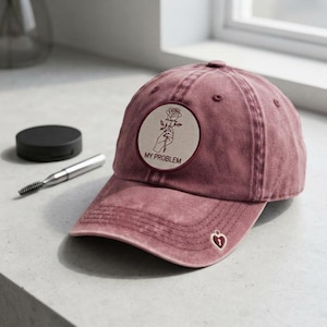 May include: A dusty rose baseball cap featuring a circular patch with a line drawing of a hand holding a rose and the text MY PROBLEM. The cap includes a small heart-shaped charm with a keyhole detail attached to the brim's edge.