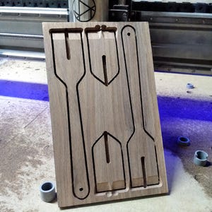 May include: A rectangular light brown wooden board featuring CNC-milled cutouts of three distinct utensil shapes. The board displays a long fork-like shape on the left, a long handle shape on the right, and two smaller shovel-like shapes in the centre.