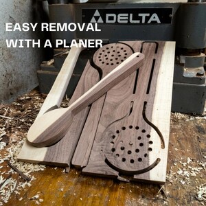 May include: A wooden board featuring cutouts of kitchen utensils, including a spoon and a slotted spoon, with the text EASY REMOVAL WITH A PLANER. The board displays natural wood grain with light and dark brown tones, positioned near a Delta machine.