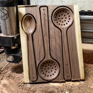 May include: A rectangular dark walnut wood board featuring three recessed kitchen utensils: a solid spoon on the left, a slotted spoon in the center, and a slotted ladle on the right. Each utensil is engraved with the name Rohrer or Laurich.