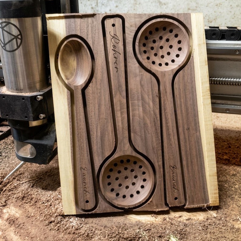 May include: A rectangular dark walnut wood board featuring three recessed kitchen utensils: a solid spoon on the left, a slotted spoon in the center, and a slotted ladle on the right. Each utensil is engraved with the name Rohrer or Laurich.