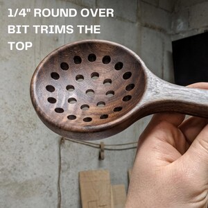 May include: A dark brown wooden slotted spoon with a circular bowl featuring multiple drilled holes. The text 1/4" ROUND OVER BIT TRIMS THE TOP is visible in the upper left corner, indicating the tool used for the edge profile of the spoon.