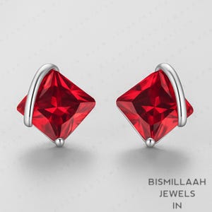 Puede incluir: Un par de pendientes de botón con piedras preciosas rojo rubí de corte cuadrado. Cada pendiente presenta un acento plateado curvado y una pequeña bola plateada en la base. El texto "BISMILLAAH JEWELS IN" es visible.
