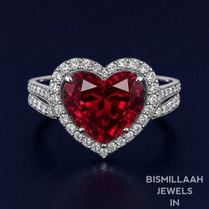 May include: A heart-shaped ruby ring with a halo of small diamonds. The ring has a silver band with additional small diamonds. The image includes the text "Bismillaah Jewels In".