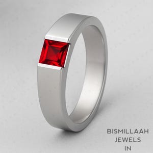 May include: A silver ring with a square, bright red gemstone. The ring has a flat band and a modern design. The gemstone is set into the band. The text "BISMILLAAH JEWELS IN" is in the bottom right corner.