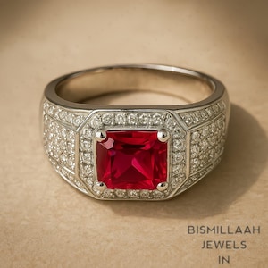 May include: A silver ring with a large, square, ruby-red gemstone at the center, surrounded by smaller, clear gemstones. The ring band is also encrusted with small, clear gemstones. The ring is set against a neutral background. The text "BISMILLAAH JEWELS IN" is visible.