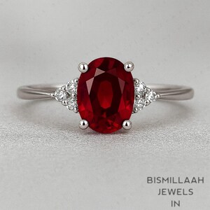 May include: A silver ring featuring a large, oval-cut, deep red gemstone as the centerpiece. Small, clear gemstones are set on either side of the main stone. The ring is set against a light gray background. The text "BISMILLAAH JEWELS IN" is visible.