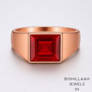 Puede incluir: Un anillo de oro rosa con una gran gema roja brillante de forma cuadrada. El anillo tiene una banda lisa y pulida y un dise&ntilde;o moderno y minimalista. La gema est&aacute; empotrada.