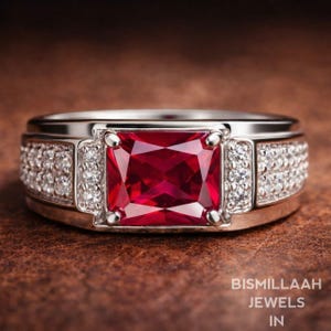Peut inclure: Bague en argent ornée d'une grande pierre rouge carrée. Des pierres plus petites et transparentes ornent les côtés de la pierre rouge. Le texte "BISMILLAAH JEWELS IN" est visible en bas.