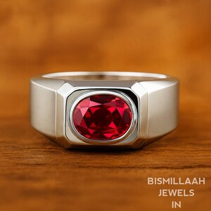 Könnte beinhalten: Ein silberner Ring mit einem großen, oval geschliffenen, tiefroten Edelstein. Der Ring hat ein breites, rechteckiges Band und eine Lünettenfassung. Der Text "BISMILLAAH JEWELS IN" ist unten rechts sichtbar.