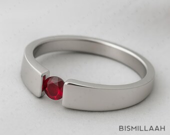 May include: A silver ring with a vibrant red gemstone. The ring has a modern design with a smooth, polished band and a bezel setting that securely holds the round ruby. The ring is a piece of jewellery.