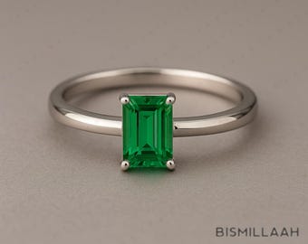 May include: A silver ring with a rectangular, emerald-cut green gemstone. The ring has a thin band and a simple, elegant design. The gemstone is a vibrant green color and is the focal point of the ring.