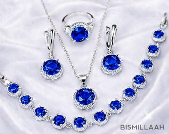 May include: A jewelry set featuring a ring, necklace, earrings, and bracelet. Each piece has a large, round, deep blue gemstone surrounded by smaller clear stones, set in silver-colored metal. The jewelry is displayed on a white, textured surface.