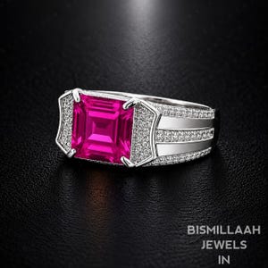 May include: A silver-toned ring featuring a large, square-cut, vibrant pink gemstone at the centre. The band is accented with rows of small, clear pavé-set stones and polished metal sections, creating a wide, structured design with a bright finish.
