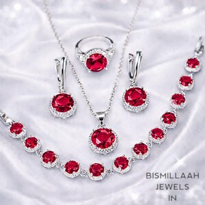 Puede incluir: Un conjunto de joyas que incluye un anillo, un collar, pendientes y una pulsera. Cada pieza tiene una gema roja grande y redonda rodeada de piedras claras más pequeñas, engastada en metal plateado. Las joyas se exhiben sobre una superficie blanca texturizada.