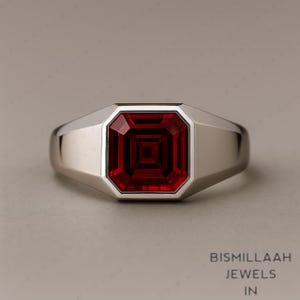 Puede incluir: Un anillo de plata con una gran gema roja oscura de forma cuadrada. La gema está engastada en un bisel y el anillo tiene un diseño simple y moderno. El anillo es probablemente un anillo para hombre.