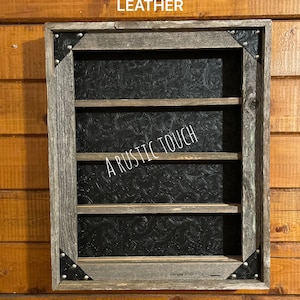 Puede incluir: Una estantería de madera rústica con tres estantes, con un respaldo de imitación de cuero negro estampado. La estantería tiene detalles metálicos plateados en las esquinas y el texto "A Rustic Touch" es visible en el respaldo.