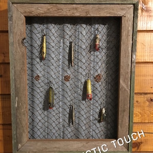 May include: A rectangular wall display featuring a weathered wooden frame surrounding a corrugated metal sheet covered with wire mesh. Seven vintage fishing lures are attached to the mesh. The text A RUSTIC TOUCH is printed in white at the bottom.