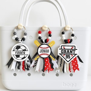 May include: White tote bag with three personalized bag charms. The charms feature a soccer ball, baseball, and baseball emblem, each with a name and number. Charms have black, white, red, and gold ribbons.