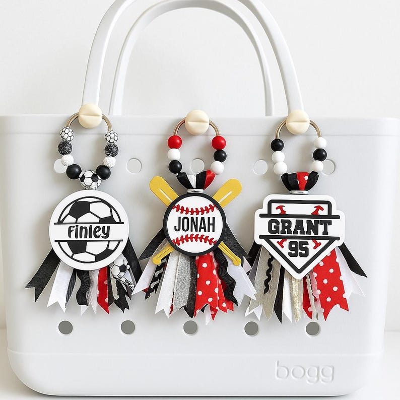 May include: White tote bag with three personalized bag charms. The charms feature a soccer ball, baseball, and baseball emblem, each with a name and number. Charms have black, white, red, and gold ribbons.