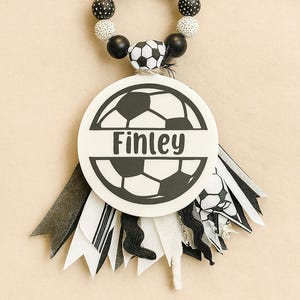 May include: A soccer-themed baby accessory featuring a white circular emblem with the name "Finley" and a soccer ball design. The accessory includes a beaded ring with black and white beads and decorative ribbons.