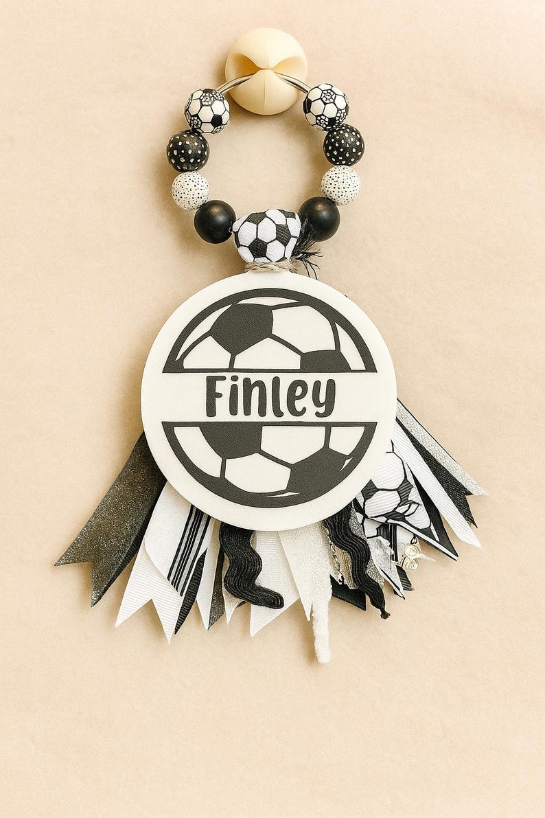May include: A soccer-themed baby accessory featuring a white circular emblem with the name "Finley" and a soccer ball design. The accessory includes a beaded ring with black and white beads and decorative ribbons.