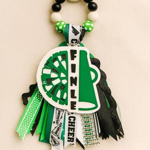 May include: A green and white cheerleading keychain with the word "FINZLE" and "CHEER" on it. The keychain has a green and white cheerleader megaphone design, black and white beads, and green and black ribbons.