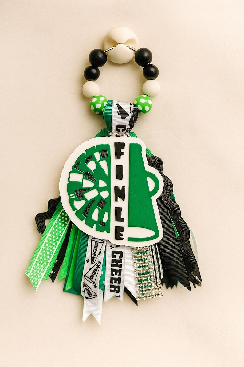 May include: A green and white cheerleading keychain with the word "FINZLE" and "CHEER" on it. The keychain has a green and white cheerleader megaphone design, black and white beads, and green and black ribbons.