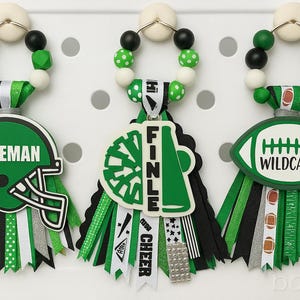 May include: Three decorative bag charms with football and cheerleading themes. Each charm features a beaded top, a ribbon base, and a themed pendant. One has a green helmet with "FREEMAN", another a megaphone with "FINLE", and the last a football with "WILDCATS".