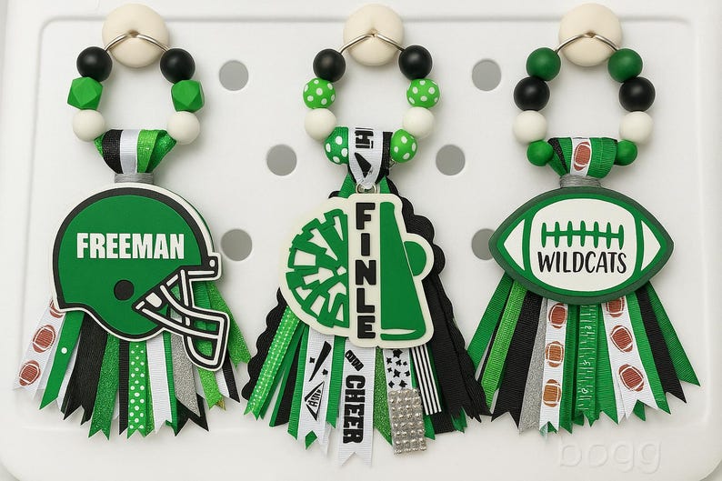 May include: Three decorative bag charms with football and cheerleading themes. Each charm features a beaded top, a ribbon base, and a themed pendant. One has a green helmet with "FREEMAN", another a megaphone with "FINLE", and the last a football with "WILDCATS".