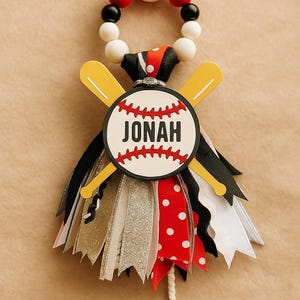 May include: A baseball-themed ornament featuring a baseball with the name "Jonah" and crossed yellow bats. The ornament has a beaded top and colorful ribbons in red, black, white, and gold. A fun decoration for a baseball fan.
