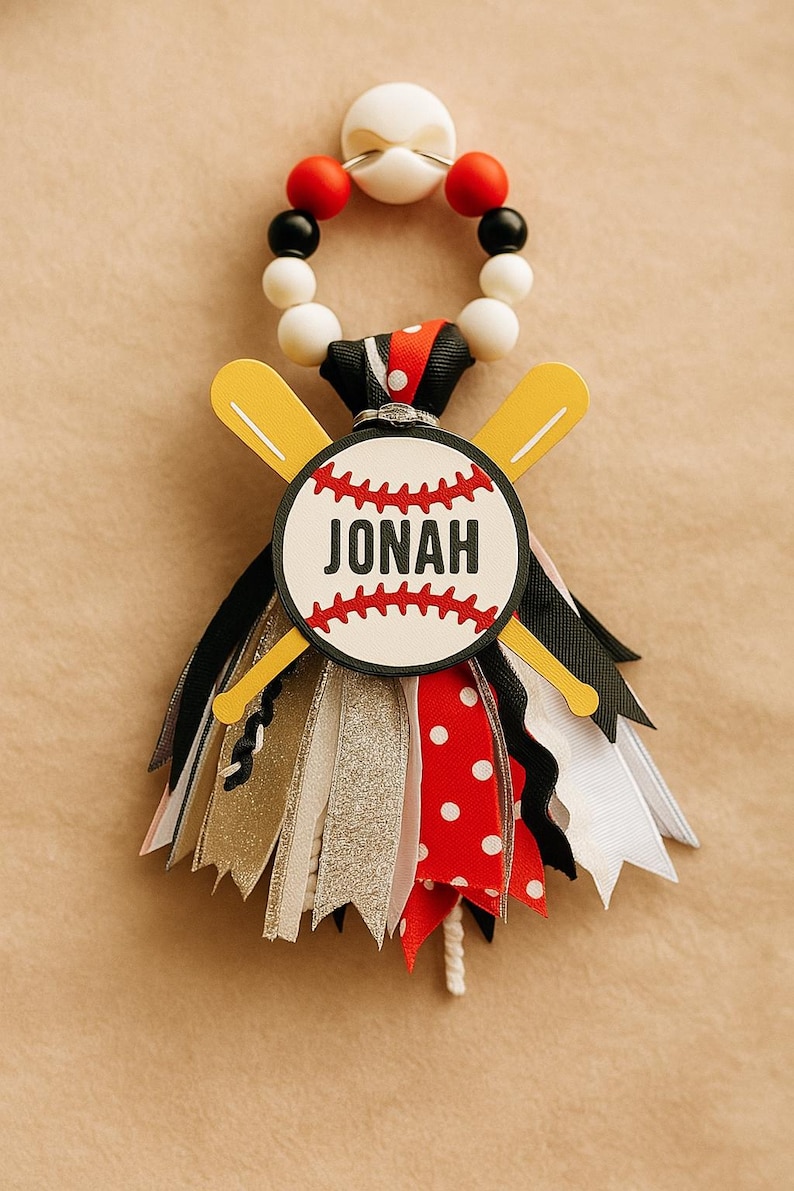 May include: A baseball-themed ornament featuring a baseball with the name "Jonah" and crossed yellow bats. The ornament has a beaded top and colorful ribbons in red, black, white, and gold. A fun decoration for a baseball fan.