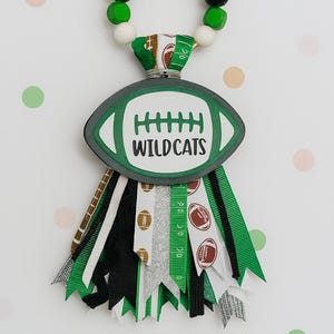 May include: A football-themed spirit item with a green and white color scheme. The design features a football shape with the word "WILDCATS" and decorative ribbons. The top has a beaded loop for hanging.