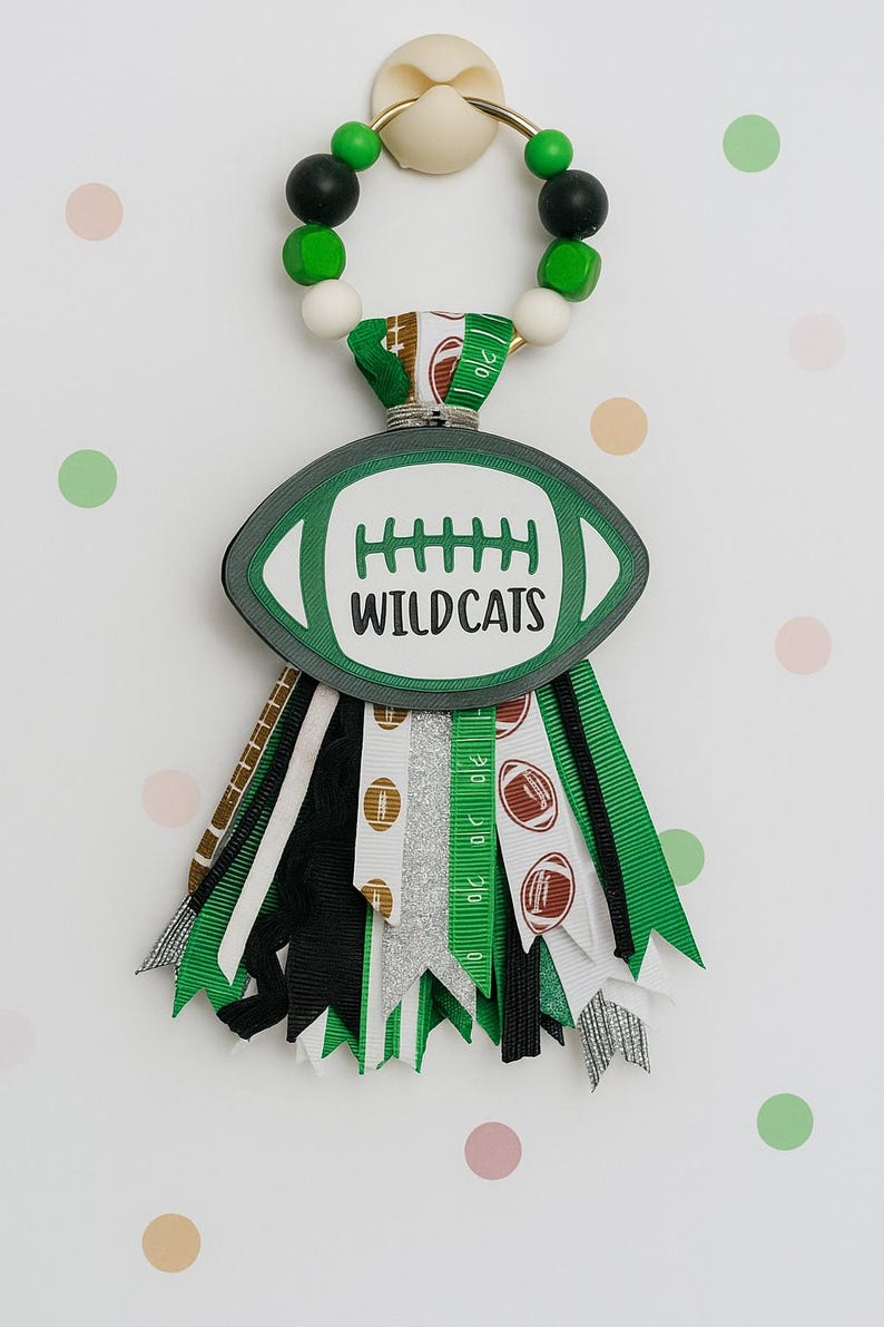 May include: A football-themed spirit item with a green and white color scheme. The design features a football shape with the word "WILDCATS" and decorative ribbons. The top has a beaded loop for hanging.