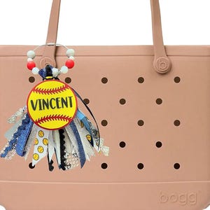 May include: A light pink tote bag with a decorative charm. The charm features a yellow softball design with the name "VINCENT" and colorful ribbons. The bag has a textured surface with black dot details.