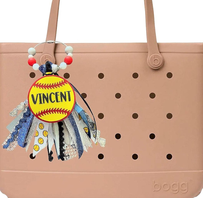 May include: A light pink tote bag with a decorative charm. The charm features a yellow softball design with the name "VINCENT" and colorful ribbons. The bag has a textured surface with black dot details.