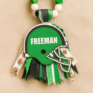 May include: A green football helmet-shaped decoration with the name "FREEMAN" in white. The decoration has a beaded loop at the top and ribbons in green, black, and white. The ribbons have football and polka dot patterns.