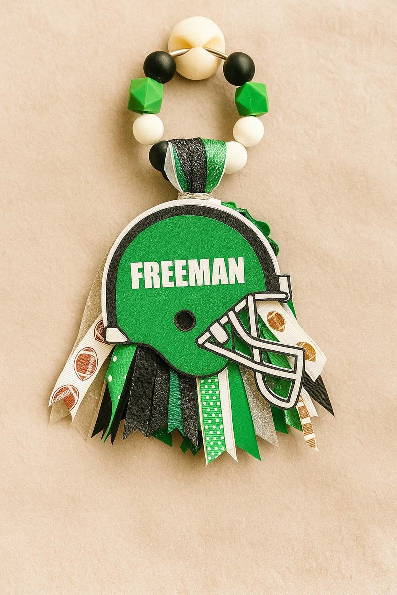 May include: A green football helmet-shaped decoration with the name "FREEMAN" in white. The decoration has a beaded loop at the top and ribbons in green, black, and white. The ribbons have football and polka dot patterns.